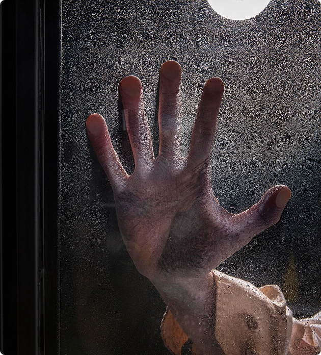 Een bleke mannenhand ligt tegen een beslagen of met druppels bedekt raam in het donker, verlicht door het licht van een lantaarn.