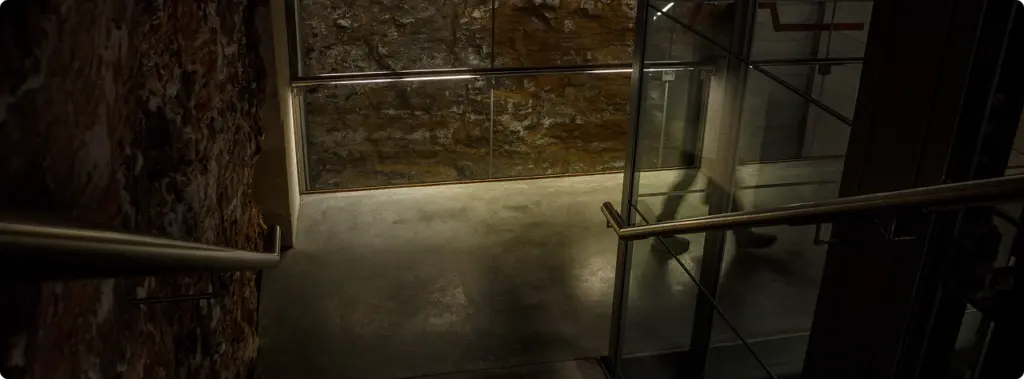 Een stenen trap die naar een ondergrondse zaal leidt, met glazen balustrades en een verlichte oude muur.