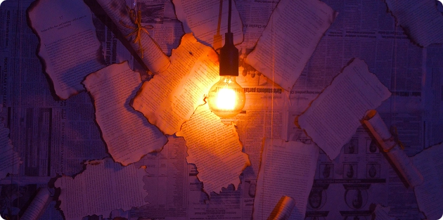 Een brandende gloeilamp werpt een paars licht op de oude, verkoolde vellen papier en rollen tegen de achtergrond van een muur van kranten.